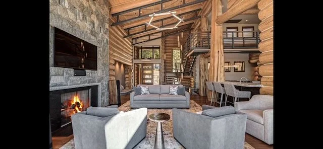 Interior of a cozy log cabin living room with gray sofas, a stone fireplace, wooden walls, and an open loft area.