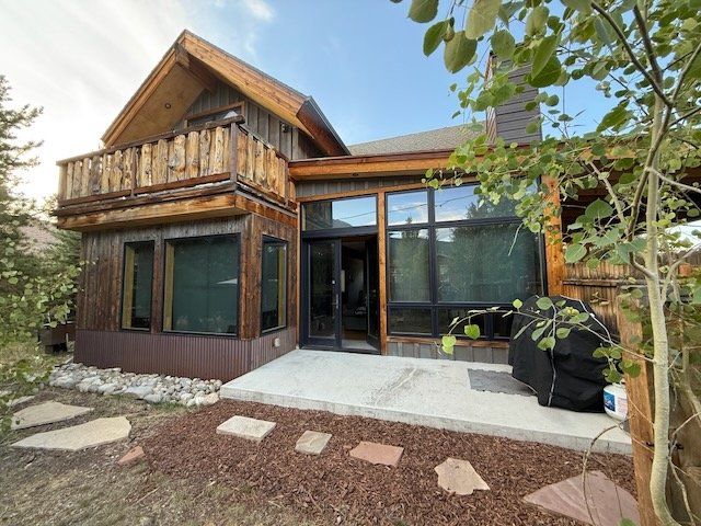 Backyard view of a two-story house with wooden siding, large glass windows, and a concrete patio with stepping stones, surrounded by trees and shrubbery.
