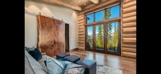 Modern living room with large windows showing outdoor forest view, wooden log walls, hardwood floors, and a gray sofa with cushions.