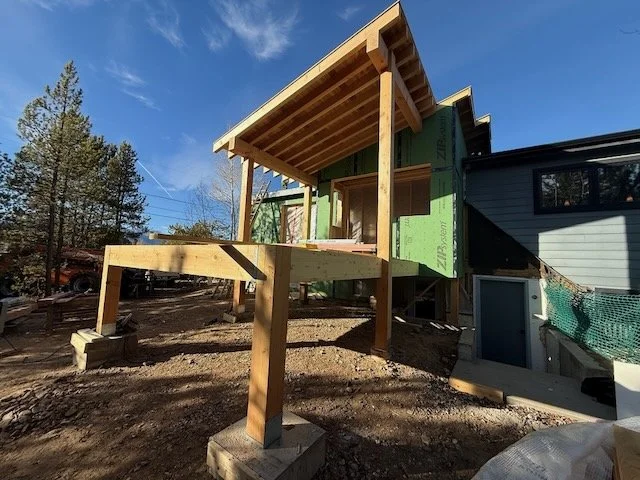 Construction site of a house with a wooden deck and roof under construction, with surrounding trees and a blue sky.