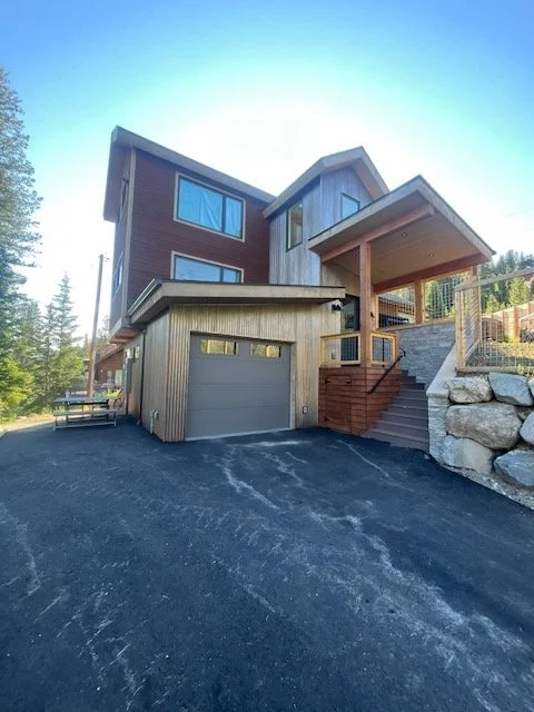 A modern house with wooden exterior siding, a garage door, and stairs leading to a front porch, set against a backdrop of trees on a sunny day.