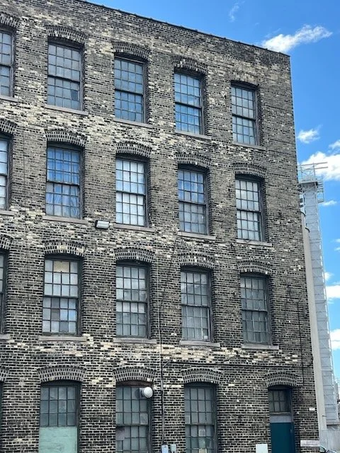 Brick building with six floors and multiple large windows, some with blinds, under a blue sky with clouds.