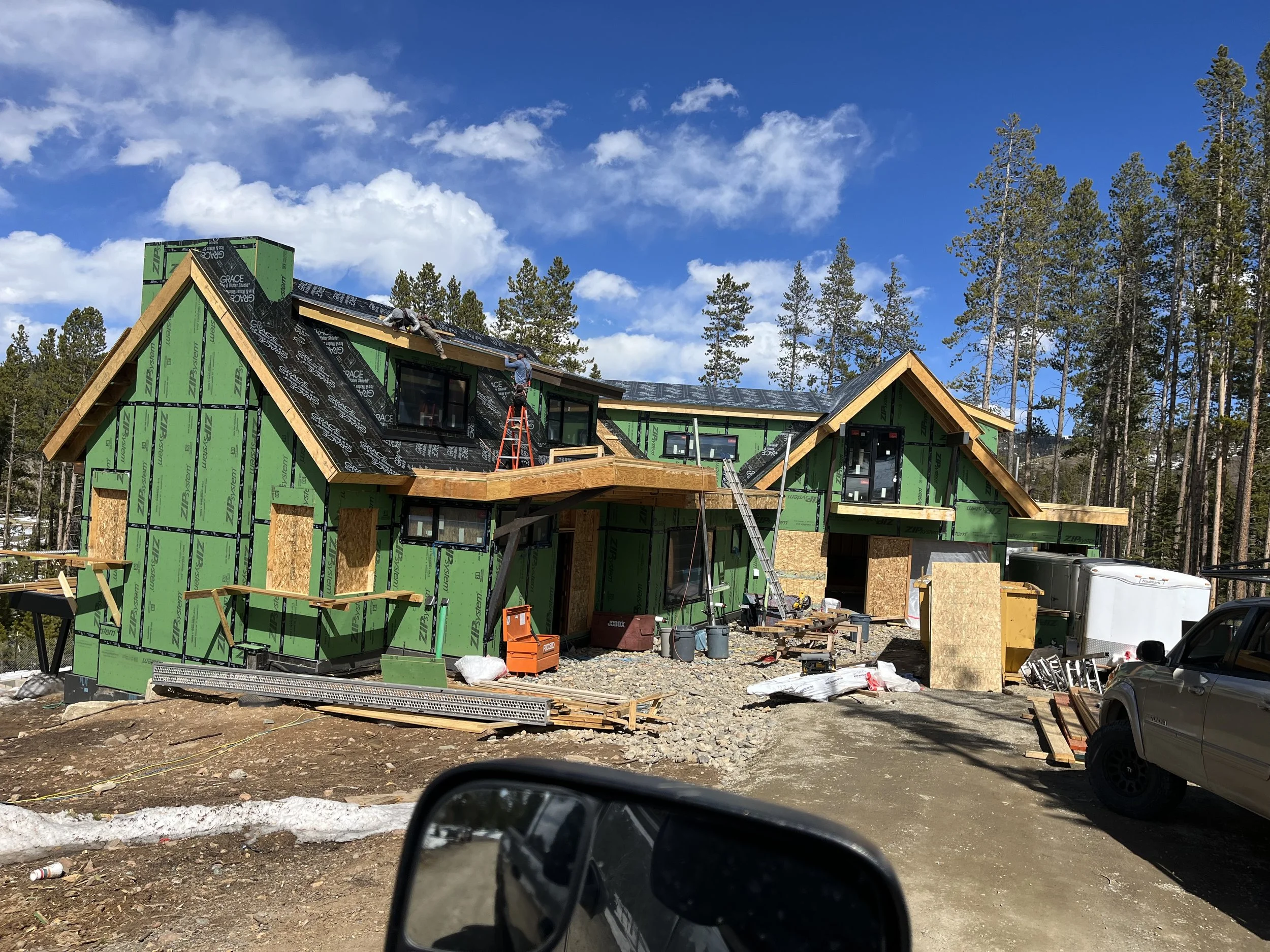 Newly under construction house with green sheathing and black roofing under blue sky, surrounded by trees.
