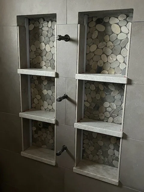 Two built-in shower niches with pebble tile backing and white shelves, separated by a central pipe in a tiled bathroom wall.