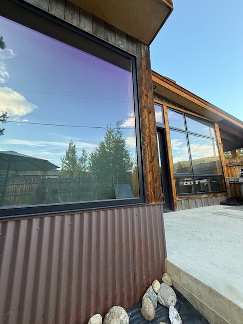 Close-up of a modern building exterior with large glass windows, wooden siding, and a concrete patio with rocks along the edge, during daytime with cloudy sky and some trees reflected in the window.