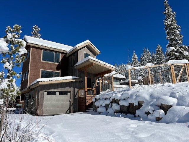 A modern two-story house covered in snow, with a garage and a wooden fence, in a snowy landscape with tall pine trees under a clear blue sky.