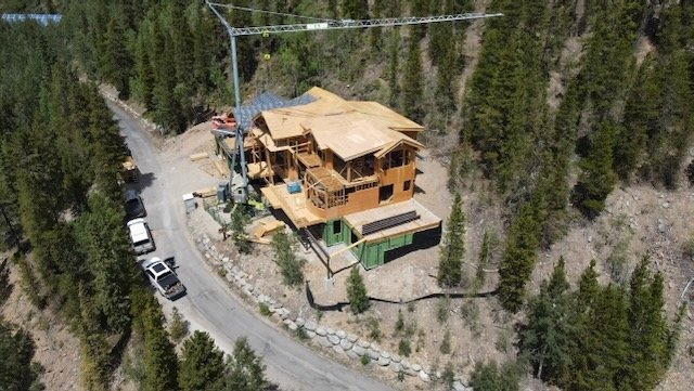 A house under construction on a hillside surrounded by trees, with construction equipment nearby and a winding road with parked cars.  Large and small projects.