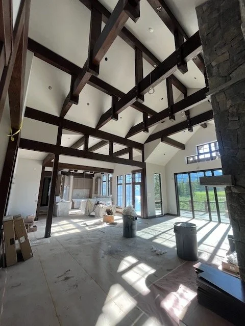 Interior view of a room under construction with exposed dark wooden beams on the ceiling, large windows, and a stone fireplace on the right, allowing natural light to fill the space.