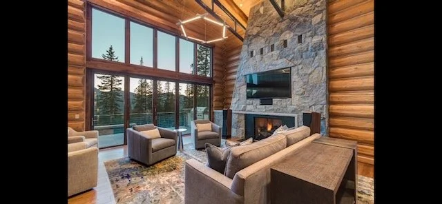 Living room with large floor-to-ceiling windows, a stone fireplace with a TV above, wood-paneled walls, and modern furniture including sofas and chairs.