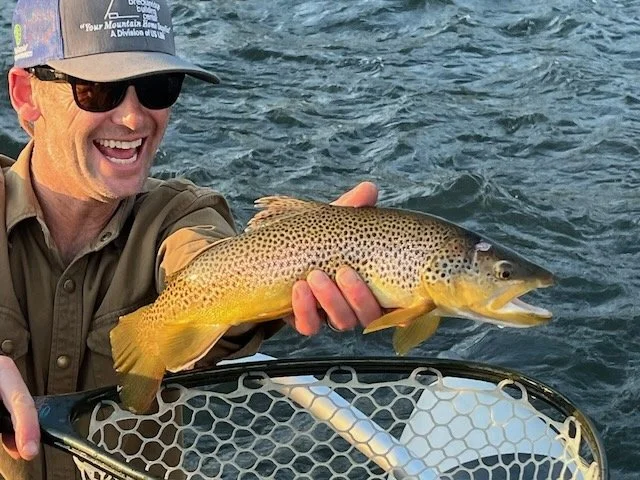 A man wearing sunglasses and a cap, smiling, holding a freshly caught fish over a body of water, with a fishing net in front of him.