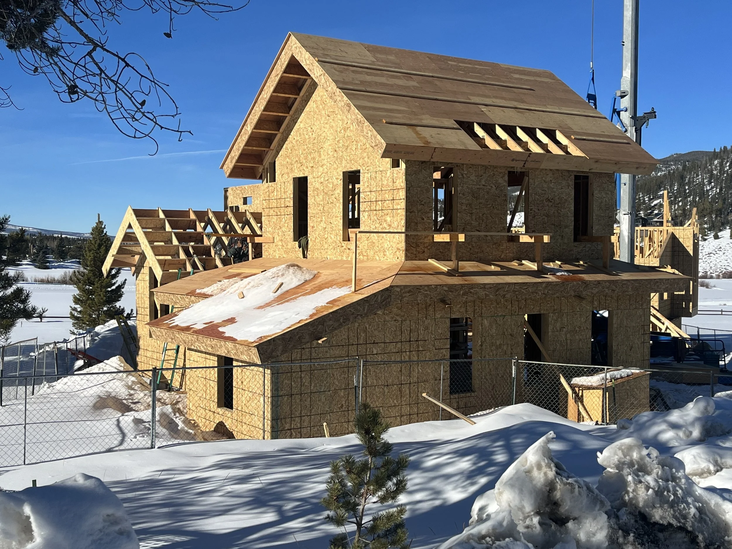 A wooden house under construction in a snowy landscape, with fenced yard, trees, mountains and a clear blue sky in the background. New Construction