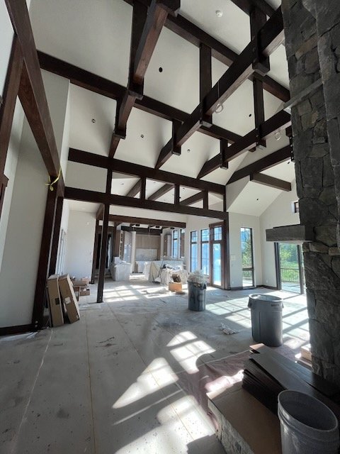 Interior of a house under construction with exposed wooden beams on the ceiling, large windows, and construction materials on the floor.