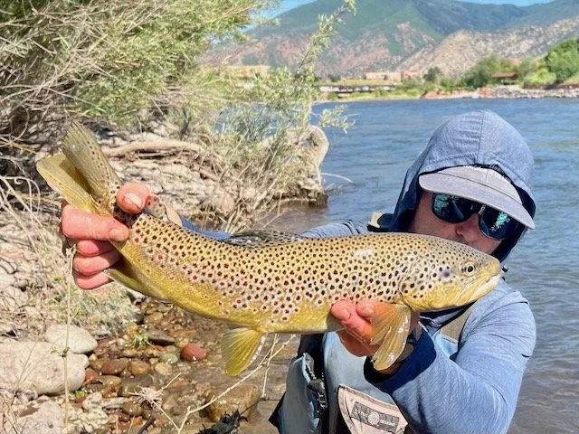 Person wearing sunglasses and a hooded jacket holding a large spotted fish near a riverbank with mountains in the background.