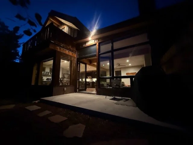 Nighttime photo of a modern house with large glass windows and wooden exterior, illuminated from inside.