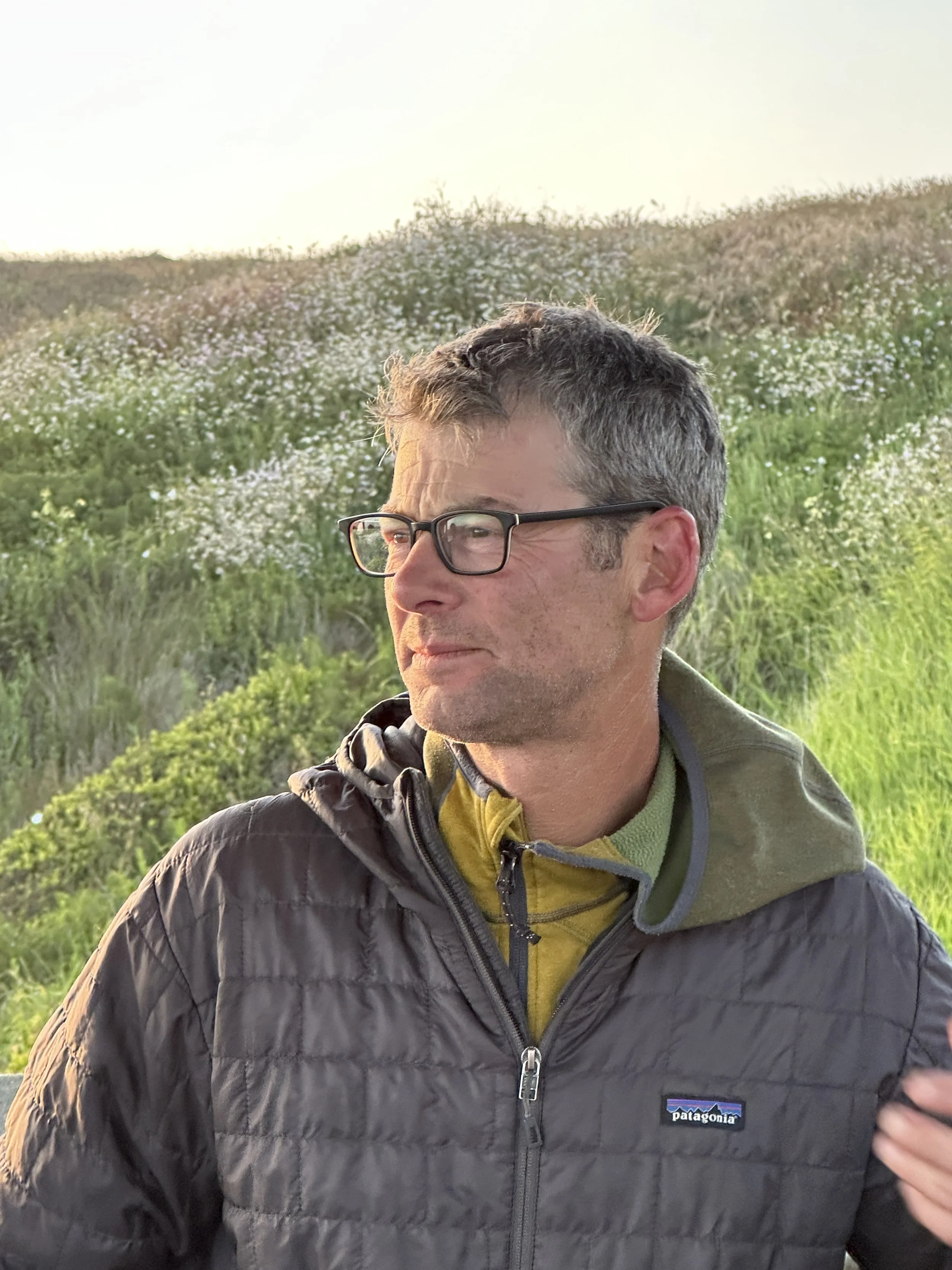 Local Professional Builder with glasses and short, dark hair stands outdoors in front of a hillside covered in flowers and greenery, wearing a black Patagonia jacket and a yellow hoodie.