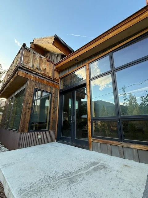 Exterior of a wooden building with large glass windows and a concrete walkway in front, set against a mountainous landscape and partly cloudy sky.