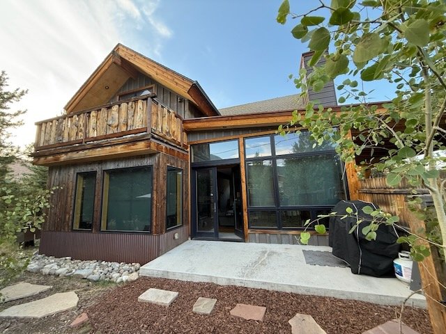 Completed a modern wooden house with large glass windows, a small balcony, and a concrete patio in a garden with a tree on the right.