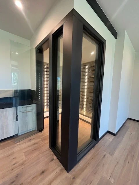 A modern wine cellar or storage room with glass doors and interior wine racks, situated in a room with light-colored walls and wooden flooring.