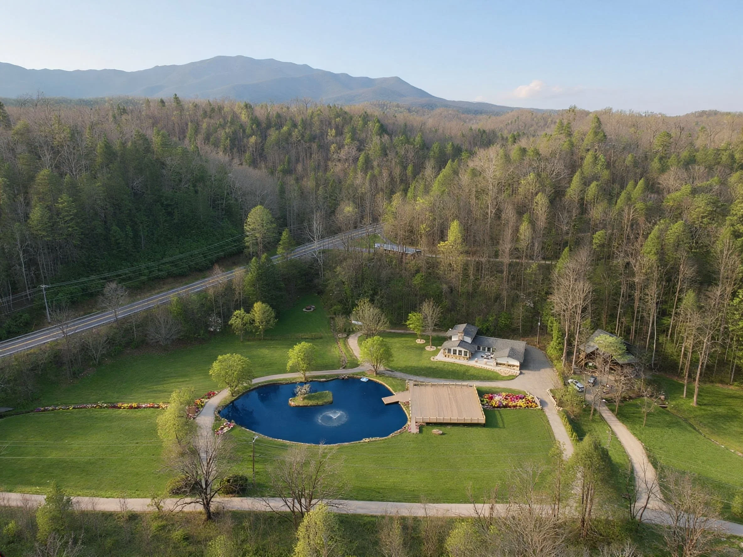 Aerial view of a large house with a pond, surrounded by green lawns, trees, and mountain backdrop.