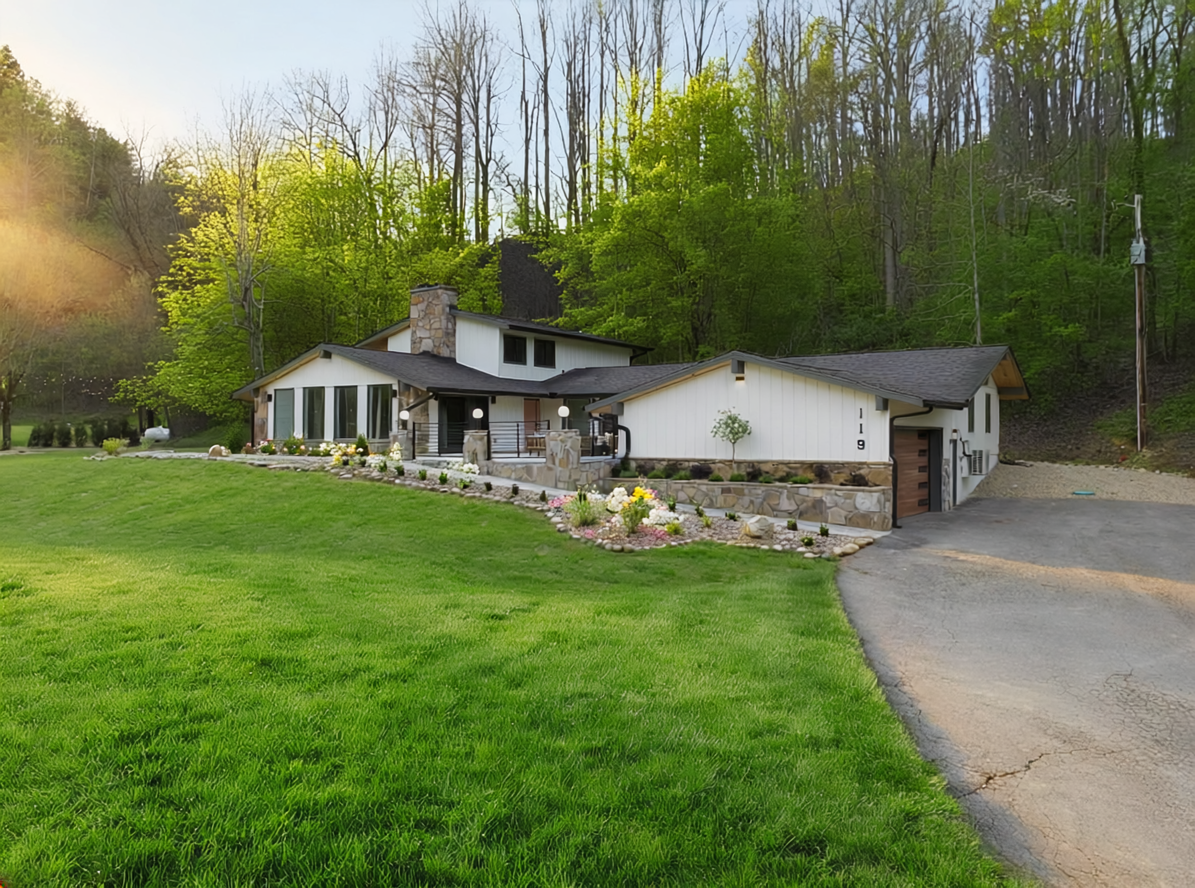 A white house with stone accents, a small front yard decorated with flowers, and a driveway leading up to a garage, situated at the edge of a wooded hill with green trees, during daylight.