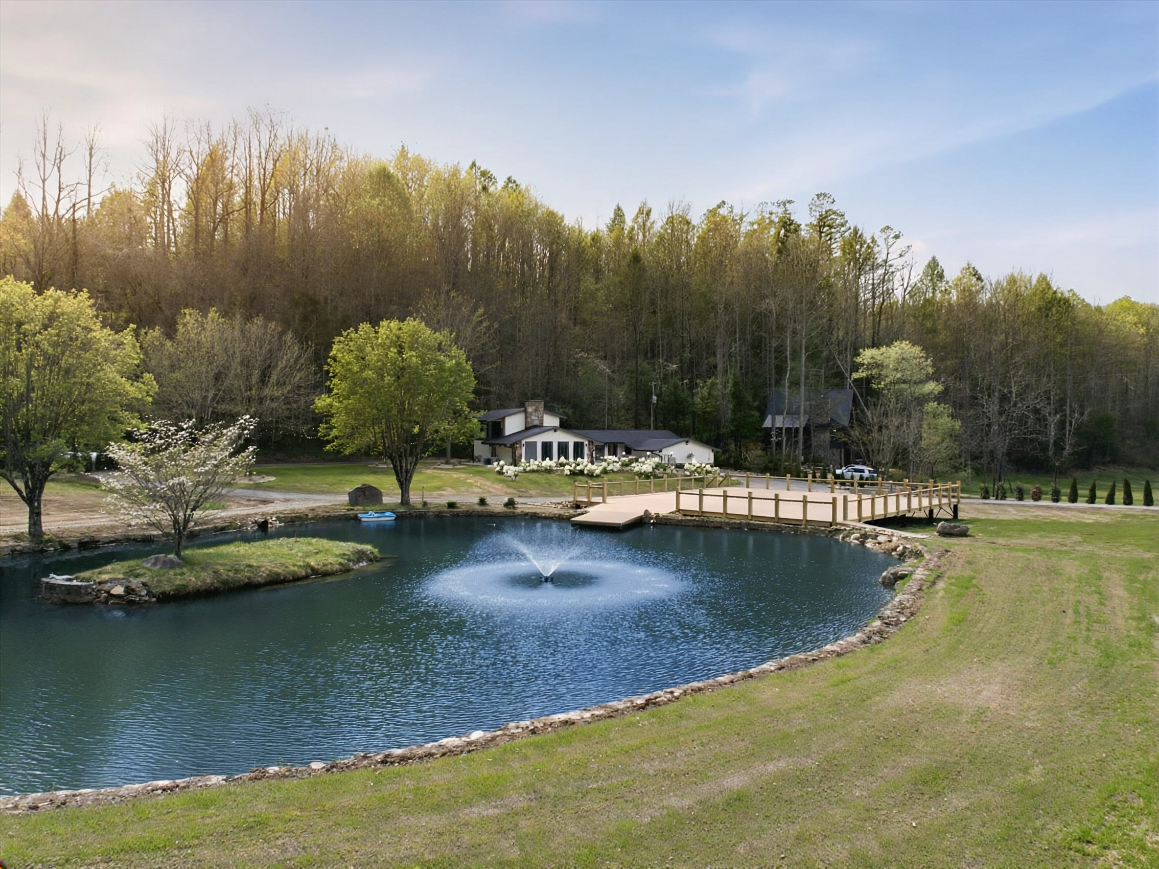 A peaceful outdoor scene featuring a small pond with a fountain, surrounded by grass and trees, and a wooden bridge leading to a modern house in the background with a wooded hillside behind it.