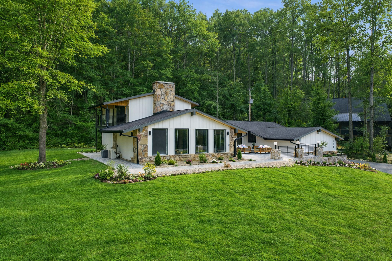 Modern house with large windows, stone fireplace, and spacious outdoor patio, surrounded by a lush green lawn and dense trees.
