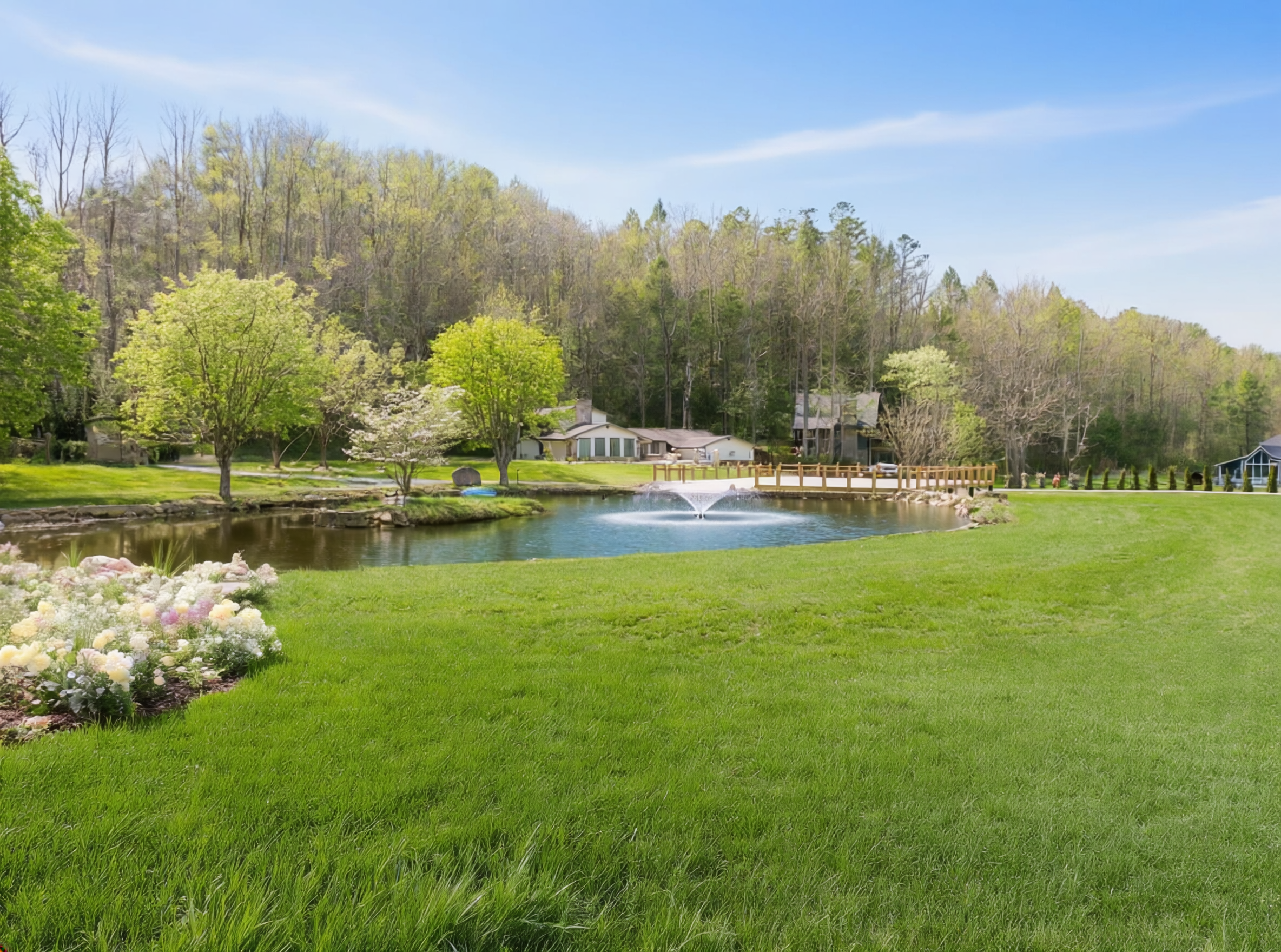 A peaceful backyard with a small pond, surrounded by green grass, blooming flowers, and trees, with houses and a wooded hillside in the background.