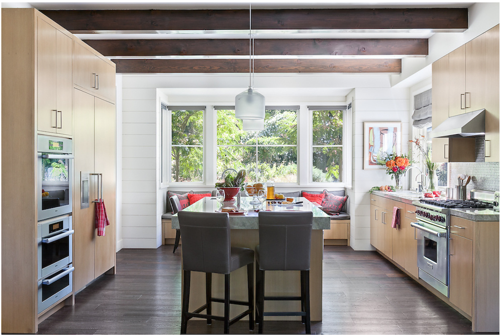 Modern kitchen with wooden cabinetry, a central island with a granite countertop, and a built-in double oven. Three black chairs are at the island. Large windows let in natural light and offer a view of green trees outside. The ceiling has exposed wo