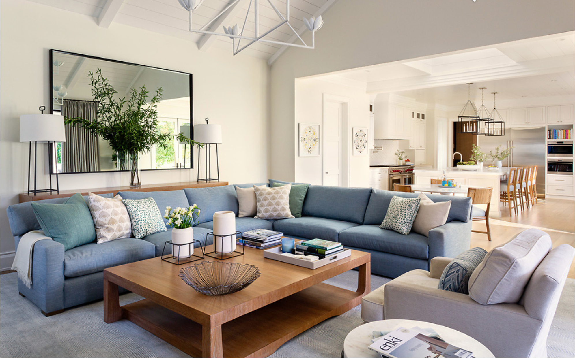 A spacious living room with a large blue sectional sofa with multiple pillows, a wooden coffee table with books and decorative objects, and a second beige armchair. There is a large mirror and two tall lamps on either side of the mirror. The backgrou