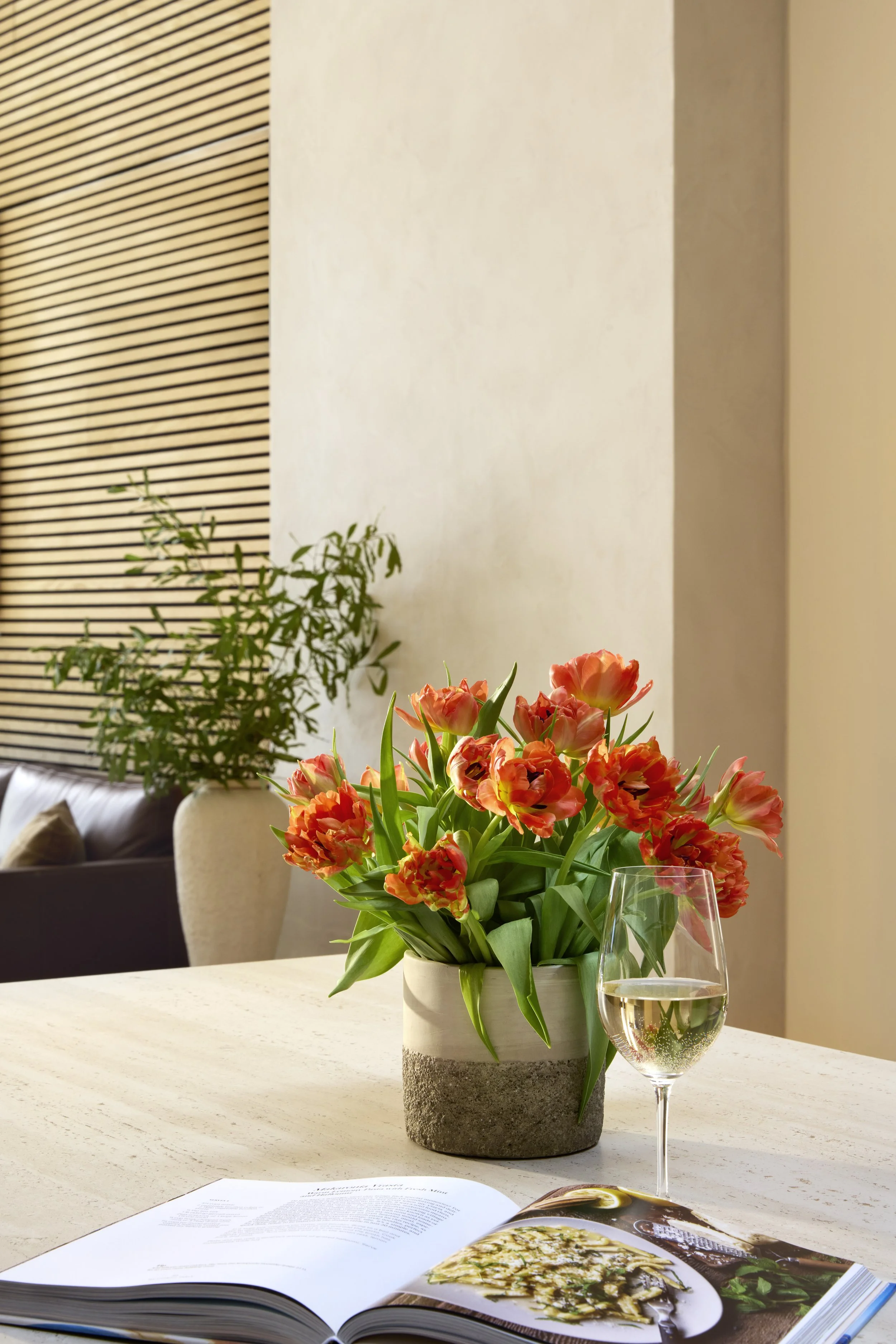 A white wall with wooden slats on the left side, a beige wall in the middle, and a part of a beige wall on the right. There is a potted plant with green foliage on the left, a bouquet of orange tulips in a speckled gray and beige vase in the center, and a glass of white wine next to the tulips. An open magazine with a food dish is on the table in the foreground.