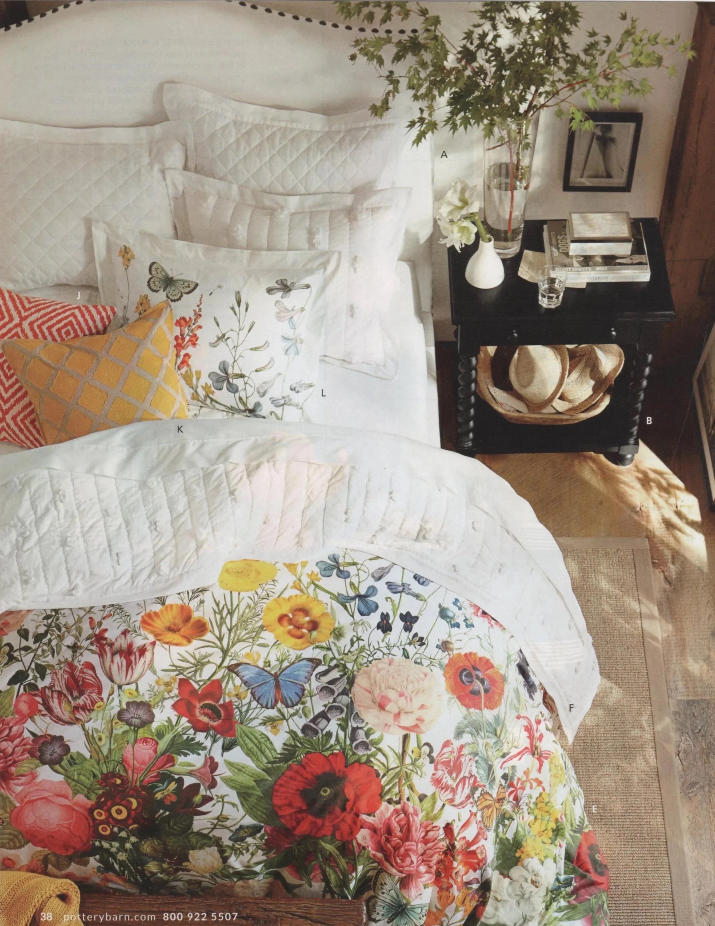 Interior Styling of a bed with white quilted bedding and decorative pillows, a black nightstand with a vase of flowers, books, and a framed picture, and a floral blanket at the foot of the bed.