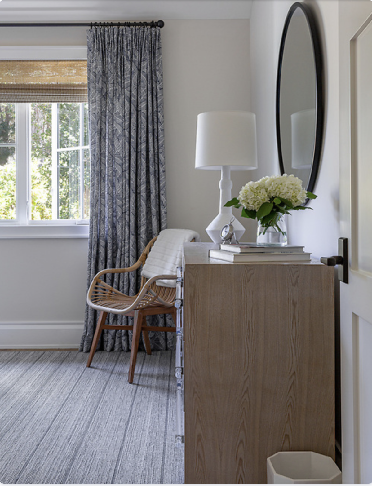 A cozy corner in a room with a window, gray patterned curtains, a light wooden dresser with a white table lamp, a vase of white hydrangeas, a stack of books, a wicker chair with a cushion, and a mirror on the wall.