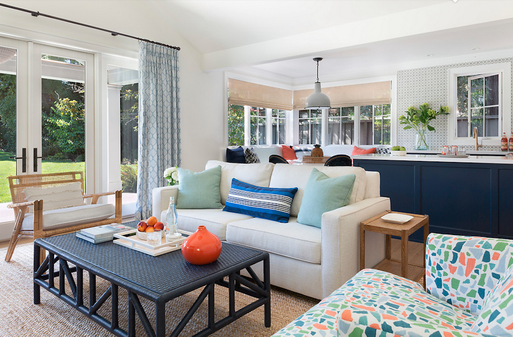 Bright living room with white sofa, patterned armchair, black coffee table, and large windows with curtains, overlooking a garden.
