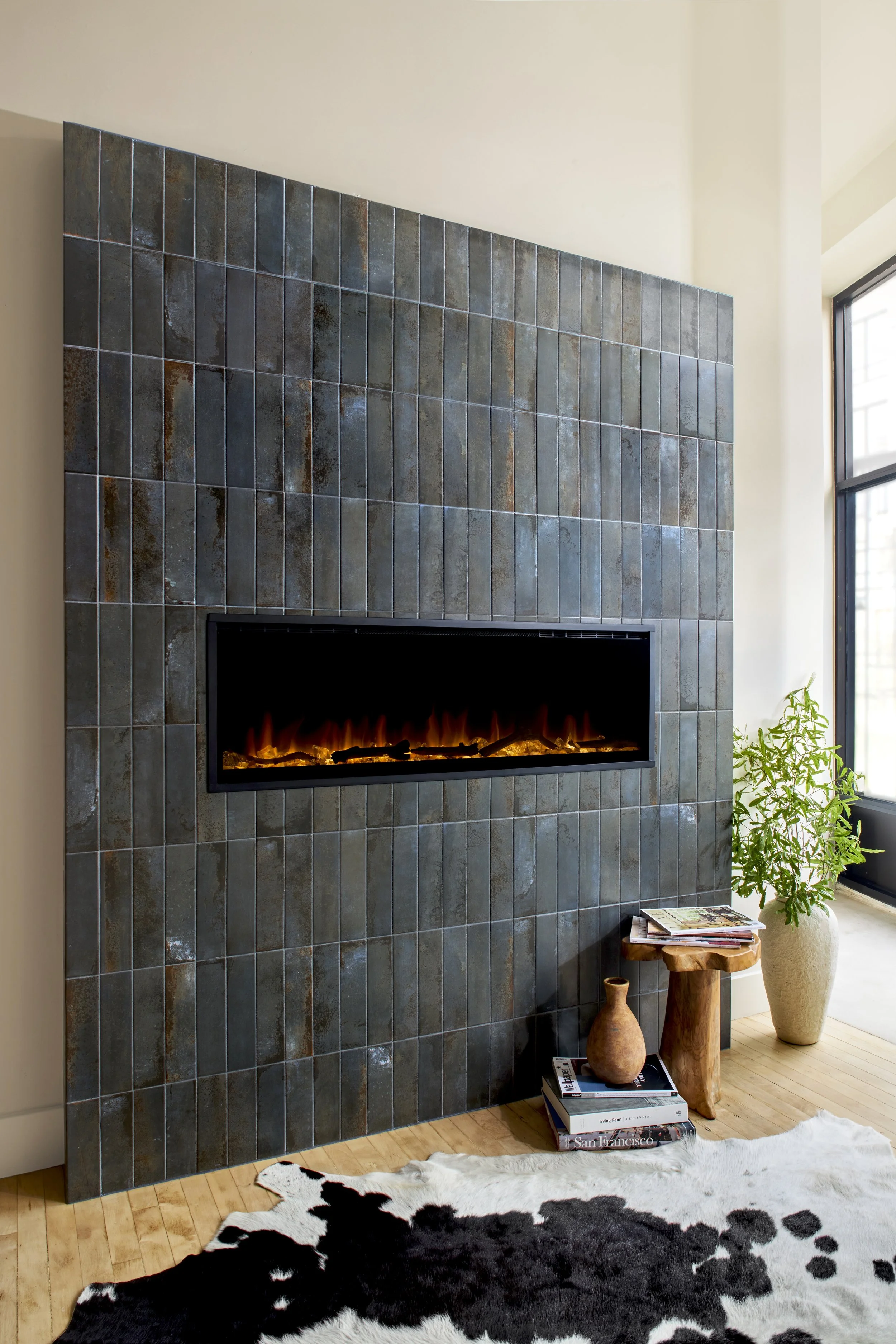 A modern living room corner featuring a built-in fireplace with stone tiles, a black and white cowhide rug on light wooden flooring, a small side table with stacked books and vases, a large vase with a leafy plant, and a large window with black framing letting in natural light.