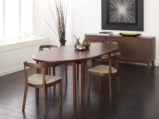 Modern dining room with an oval wooden table, four matching chairs with cushioned seats, a sideboard, and decorative vases on the table. There is a framed artwork and a bowl of green apples on the sideboard, with a tall vase and dried branches nearby
