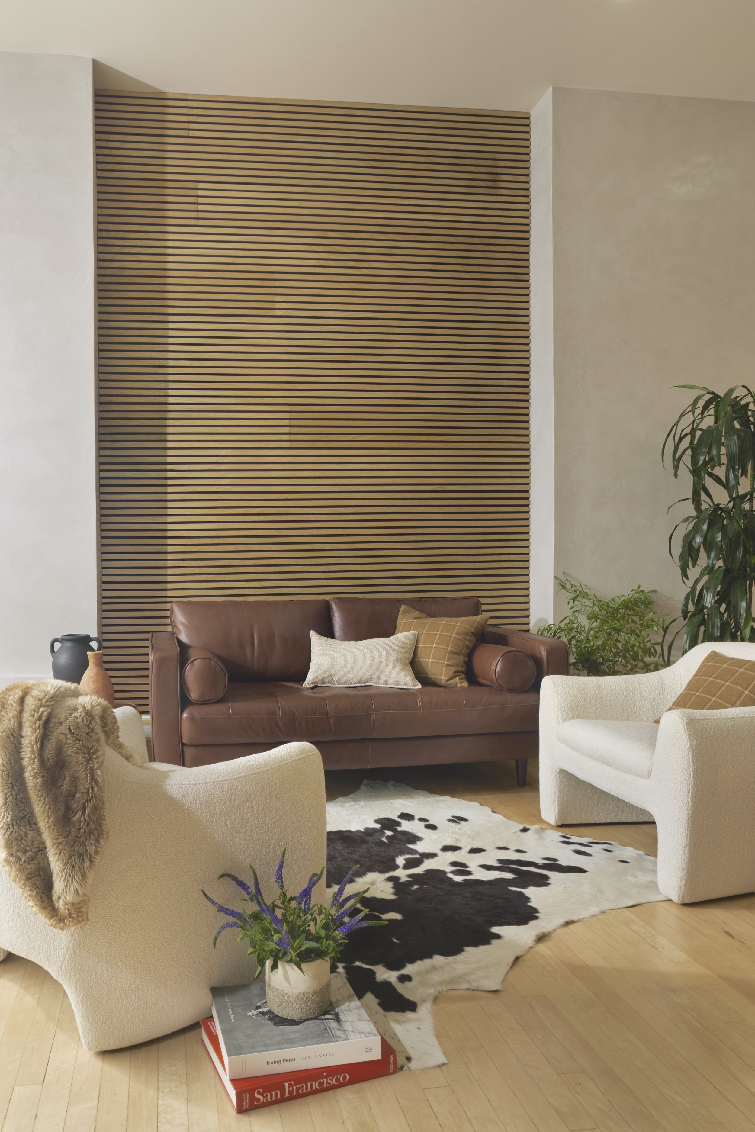 Interior living room with beige and white furniture, a black and white cowhide rug, a wooden floor, a large plant, and a wooden slat wall.