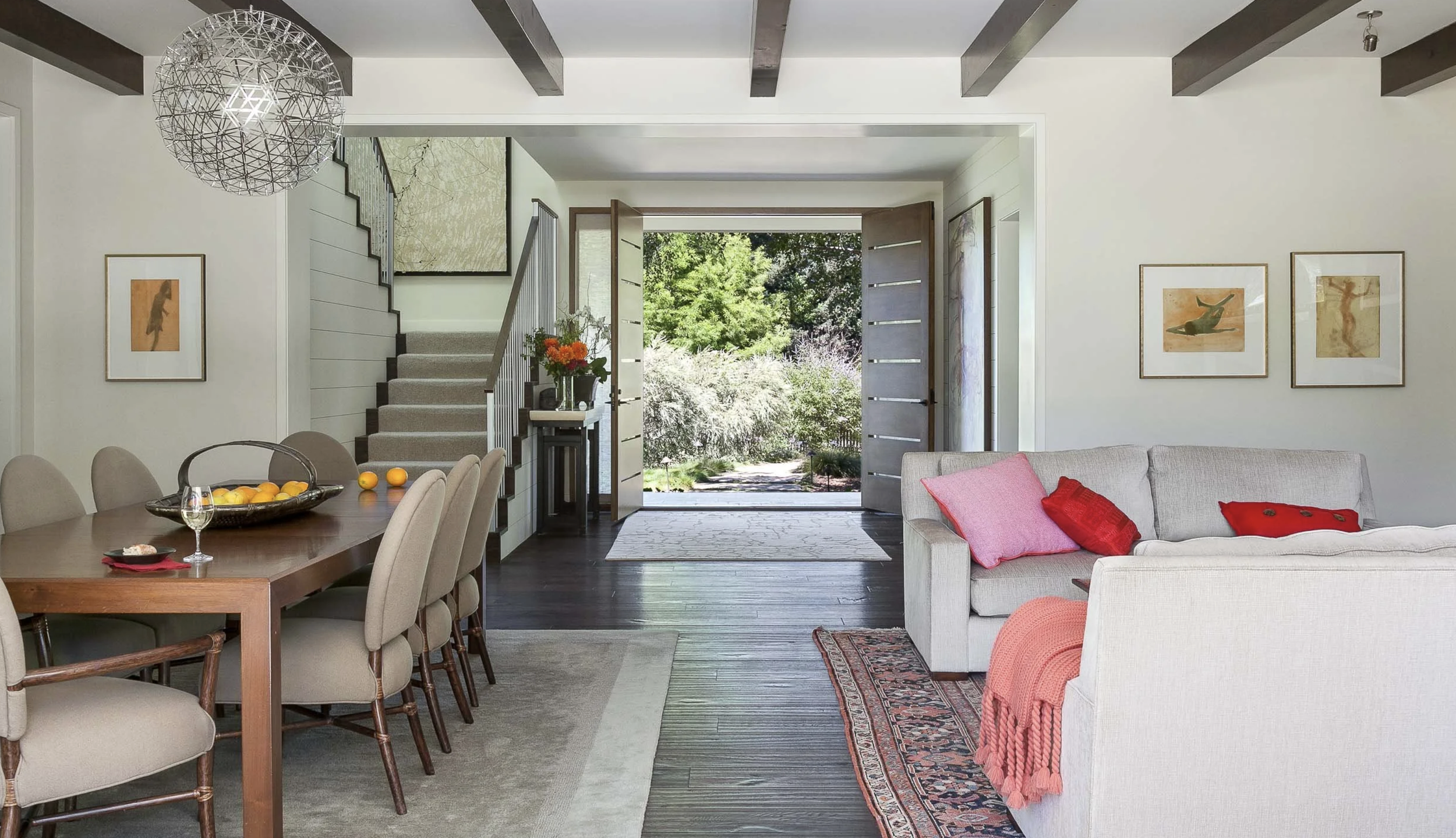 A spacious, modern living room with an open front door leading to a garden. The room features a dining table with beige chairs, a chandelier, a beige sofa with red pillows, and framed art on the walls. A side table holds a vase with flowers, and natu