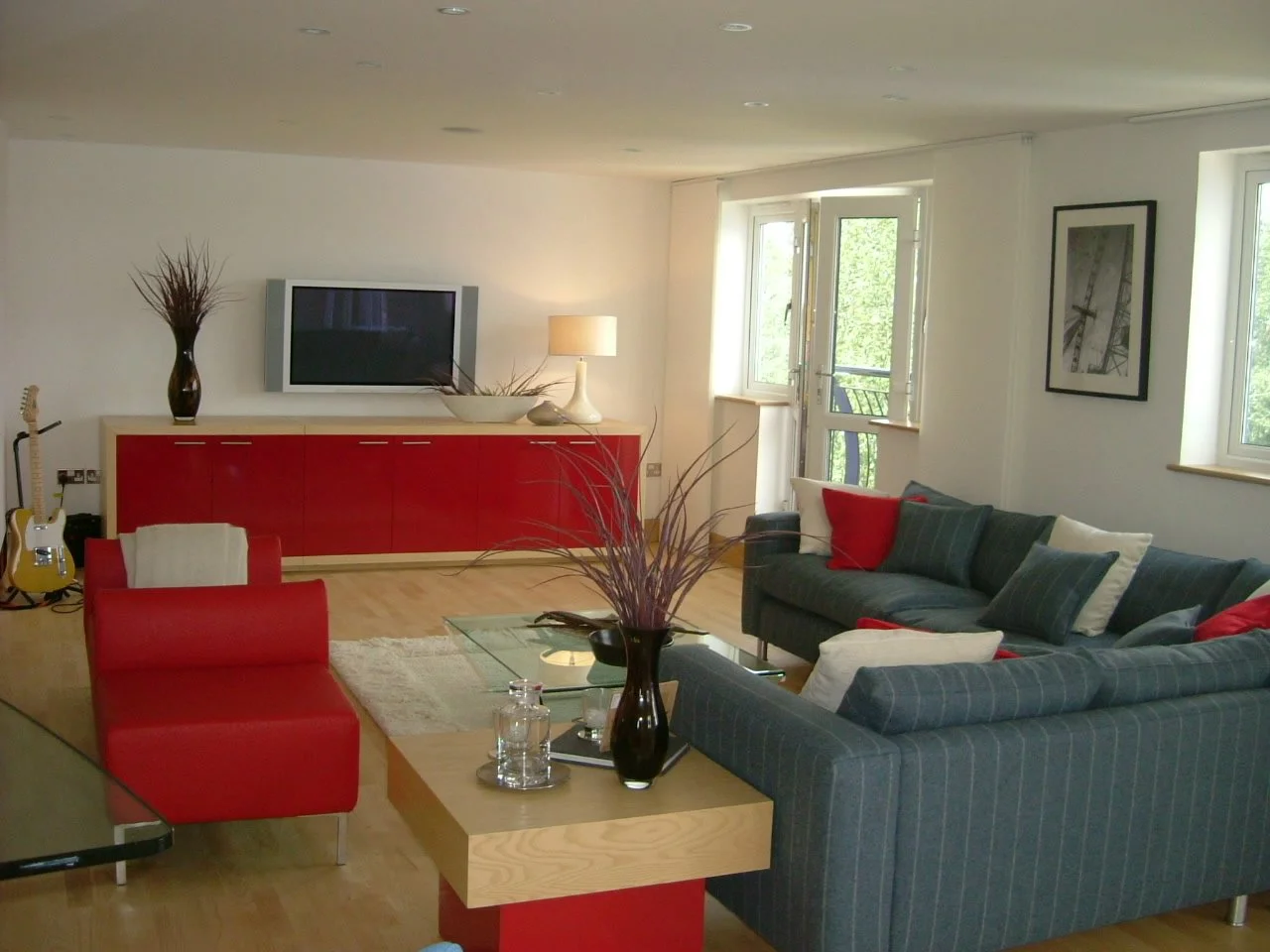Living room with a dark green sofa, red armchair, wooden coffee table with a vase of dried plants, TV on a red cabinet, guitar in corner, floor lamp, framed black and white photo on wall, windows with green trees outside.