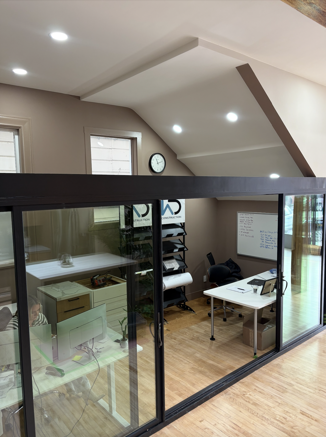 Interior view of an office space with glass walls, a whiteboard, a desk, chairs, and a staircase, with windows and a clock on the wall.