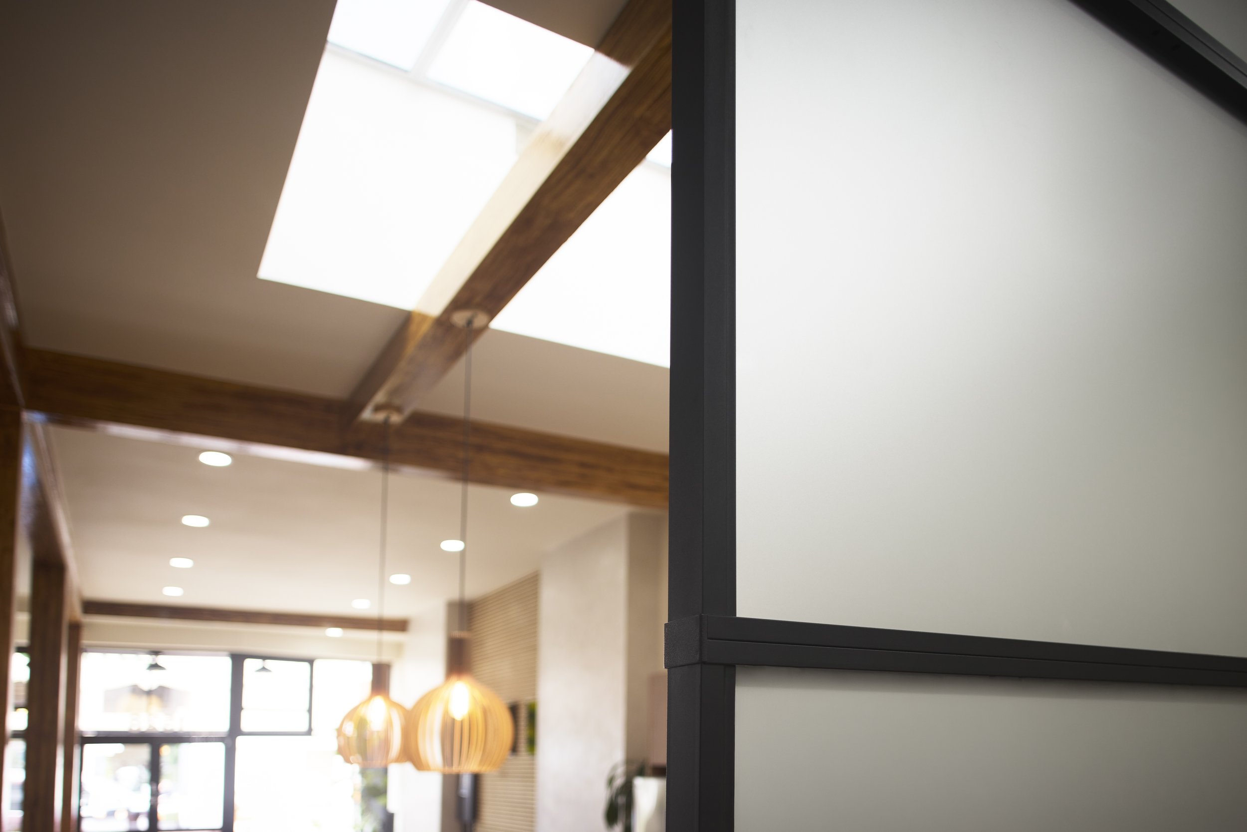 Interior view of a modern house with a skylight ceiling, wooden beams, ceiling lights, and hanging pendant lights, taken partially through a black-framed partition.