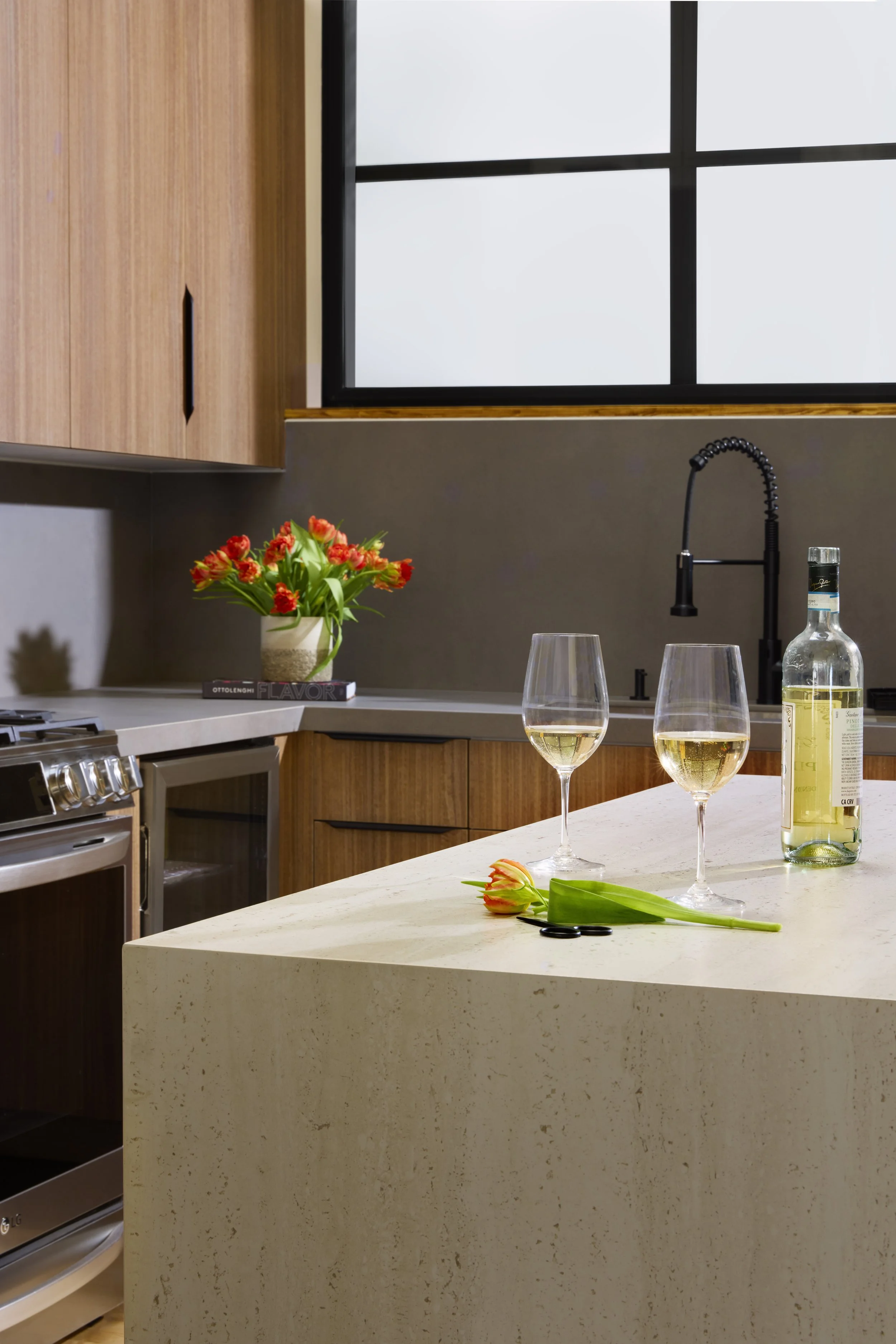 A modern kitchen with a beige quartz countertop, a black faucet, two wine glasses filled with white wine, a green tulip with orange tips, a closed pair of scissors, a bottle of white wine, a potted plant with red and orange flowers, and a window with a black frame and wooden trim.