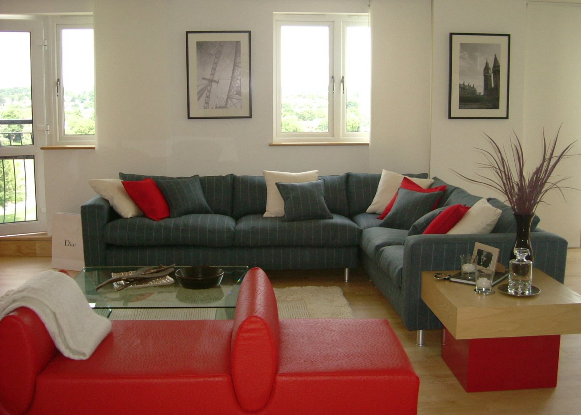 Interior Design ,concept to Installation ; Living room with a grey pin striped sofa adorned with red and white pillows, a glass coffee table with decorative items, and framed artwork on the walls. Large windows offer a view of greenery outside.