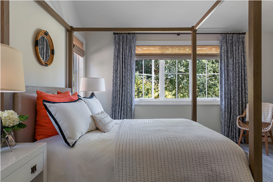 A cozy bedroom with a four-poster bed, white bedding, red and white pillows, and a neutral-toned headboard. There is a nightstand with a lamp and flowers, a mirror on the wall, large windows with blue patterned curtains, and a chair in the corner.