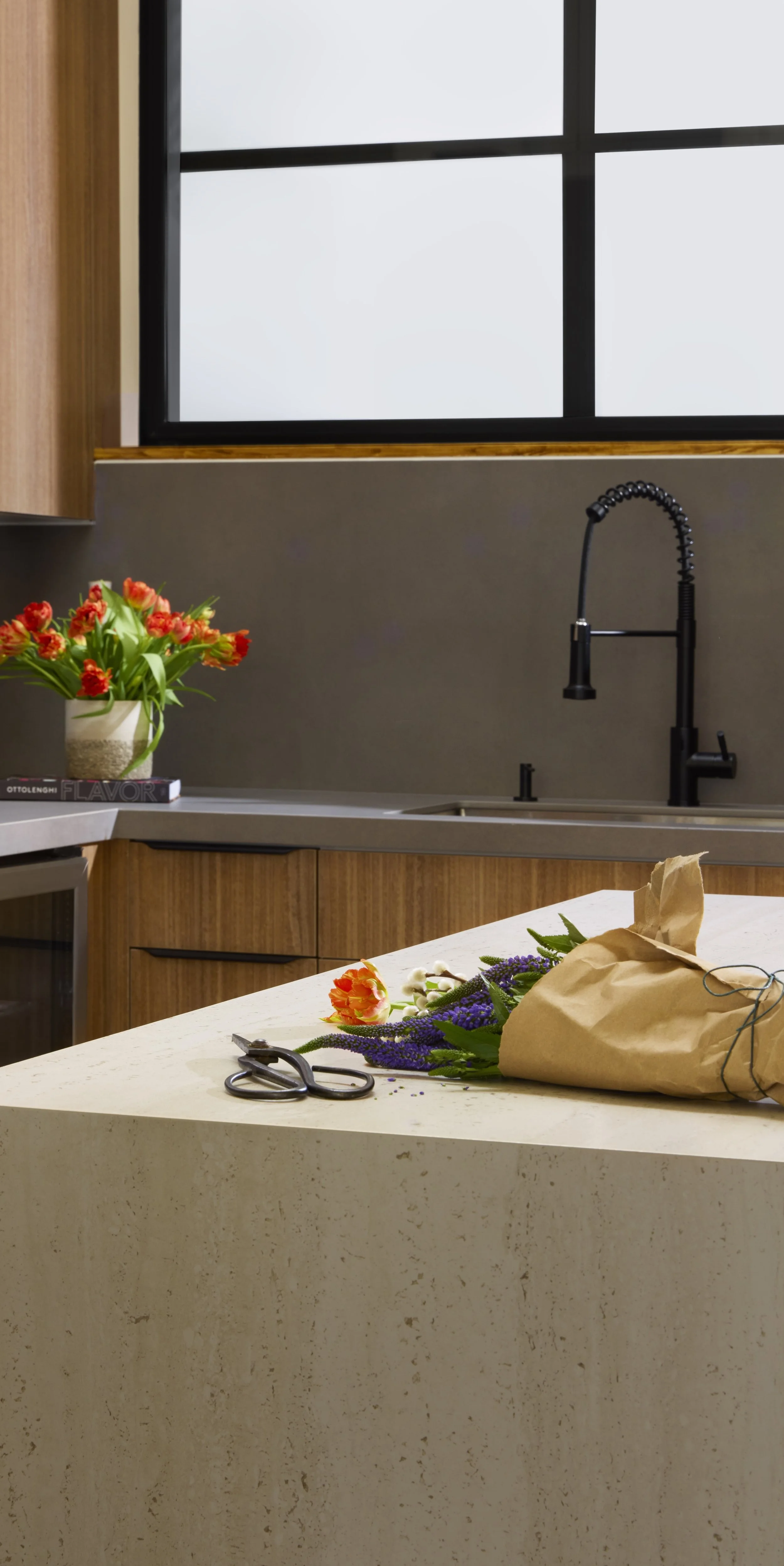 A kitchen with a countertop, flowers, scissors, and a paper bag, with a window and sink in the background.