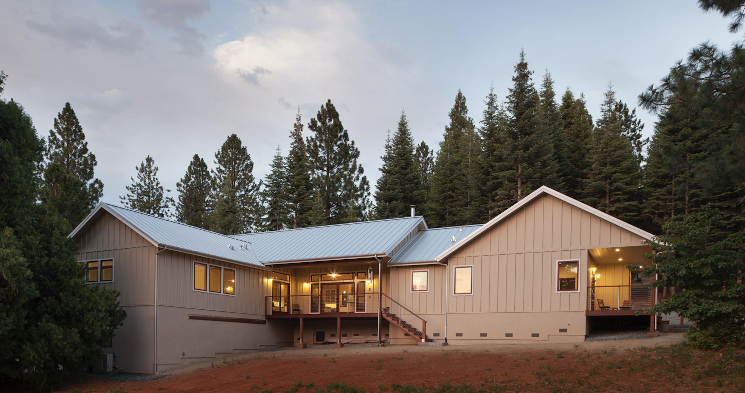 Modern house with beige siding and large windows, surrounded by tall pine trees, during dusk with interior lights on.
