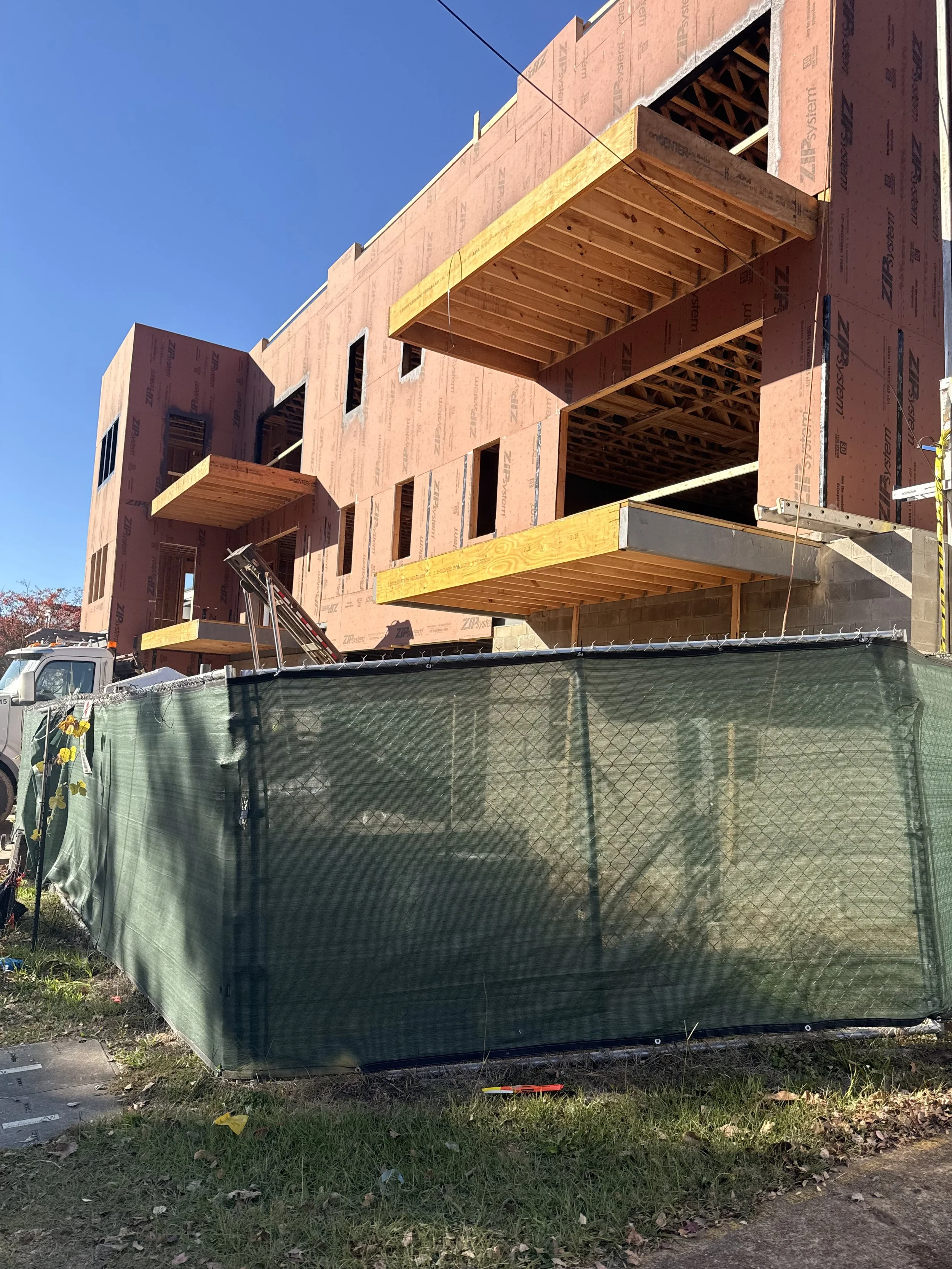Construction site with a multi-story building under construction, pink sheathing, and wooden balconies, partially obscured by green fencing.