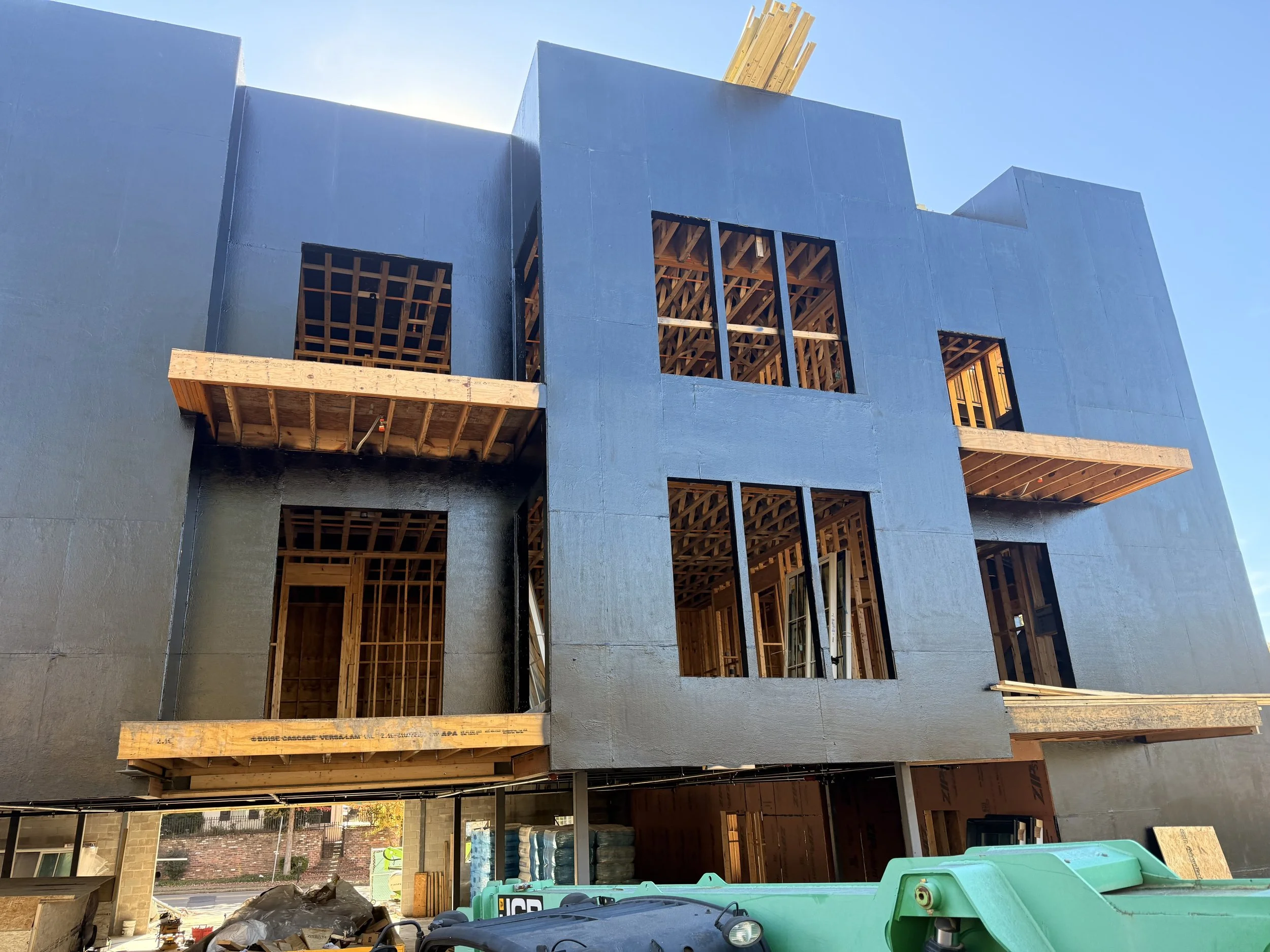 Under construction multi-story building with exposed wooden framework and exterior sheathing, with construction materials and equipment at the ground level.