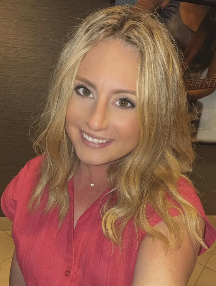 A smiling woman with blonde wavy hair wearing a pink shirt, posing indoors.
