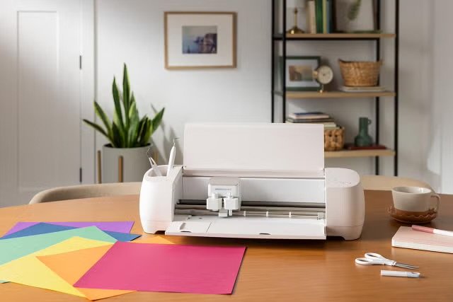Silhouette of a cricut cutting machine on a wooden table with colorful paper sheets, a copper-colored mug, a pair of scissors, and a marker, in a cozy home office with a bookshelf and plant in the background.