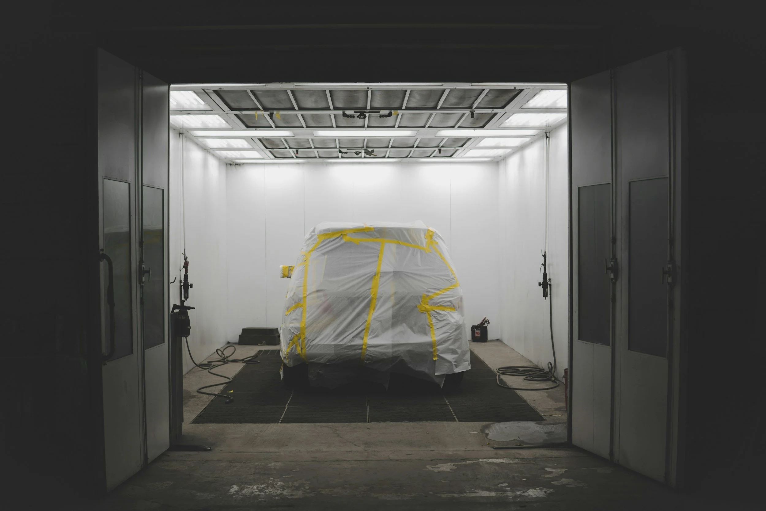 A car covered in white plastic and yellow tape inside a well-lit spray booth.
